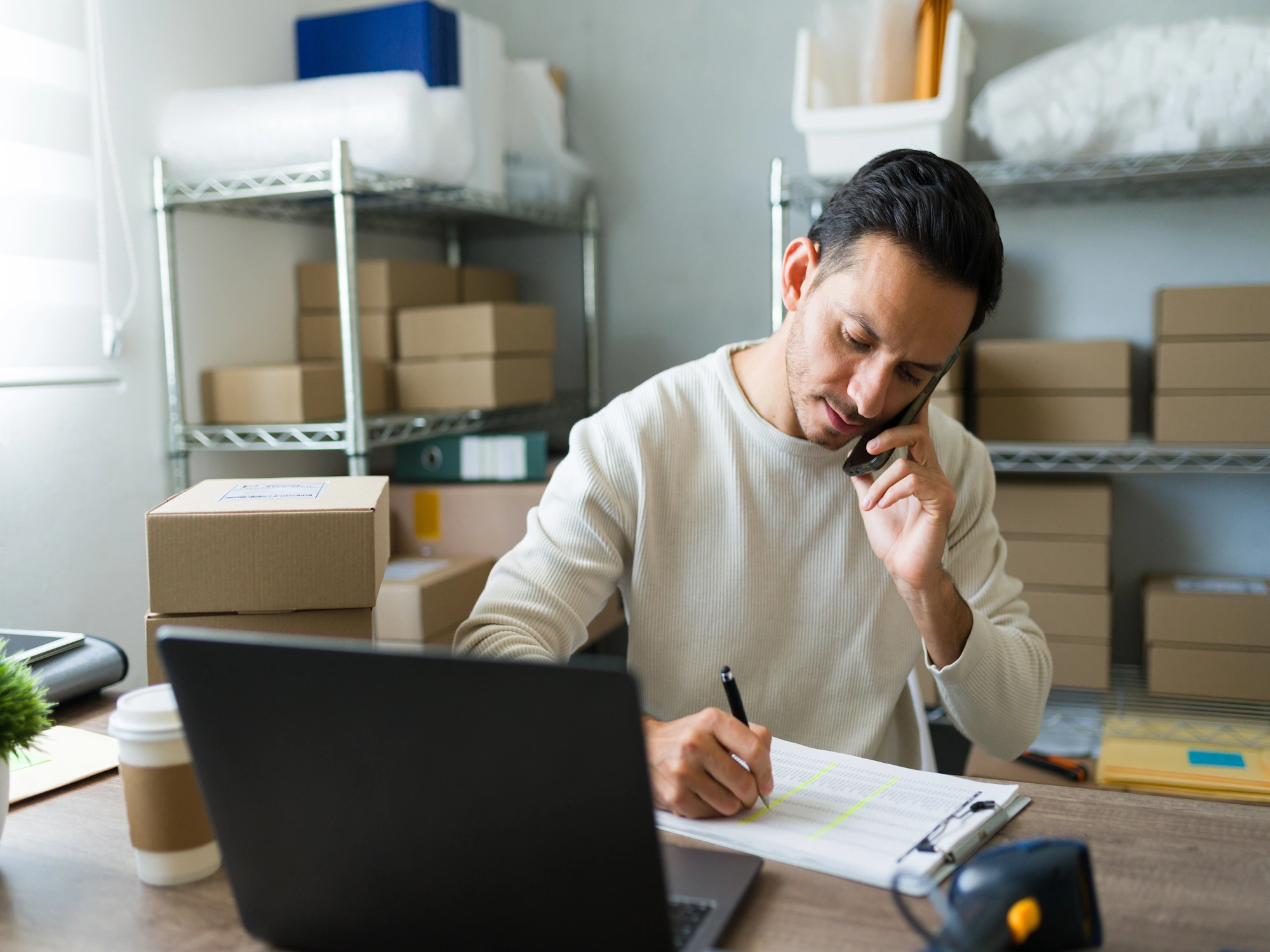 A man on the phone, writing on a clipboard next to a laptop, with shelves of boxes in the background, suggesting customer communication.