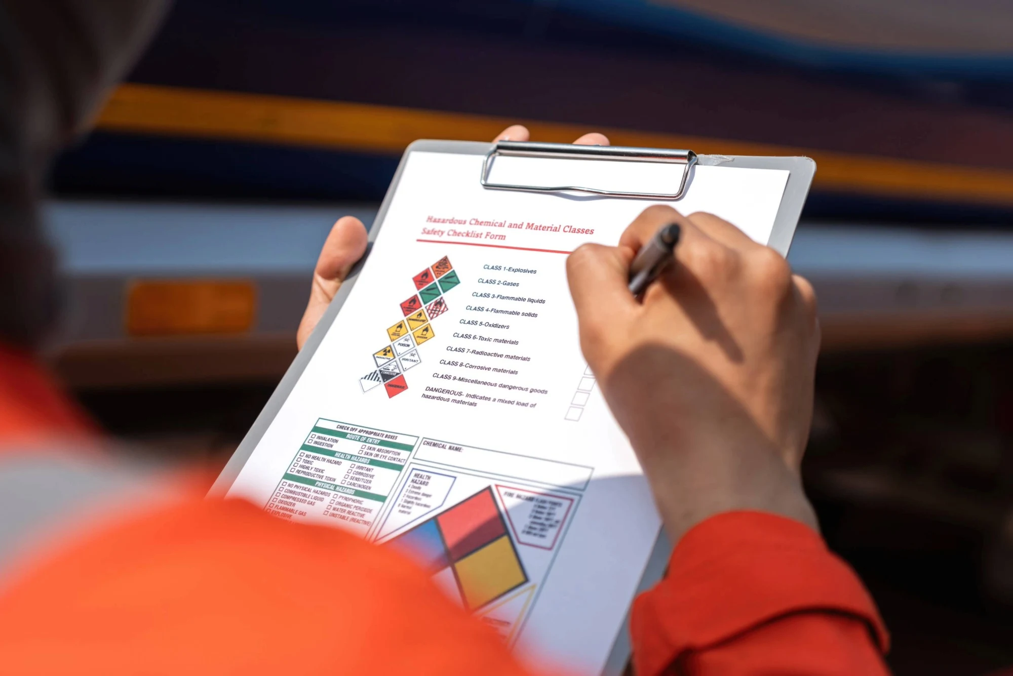 A worker in high-visibility orange clothing fills out a ‘hazardous chemical and material classes safety checklist’ on a clipboard.